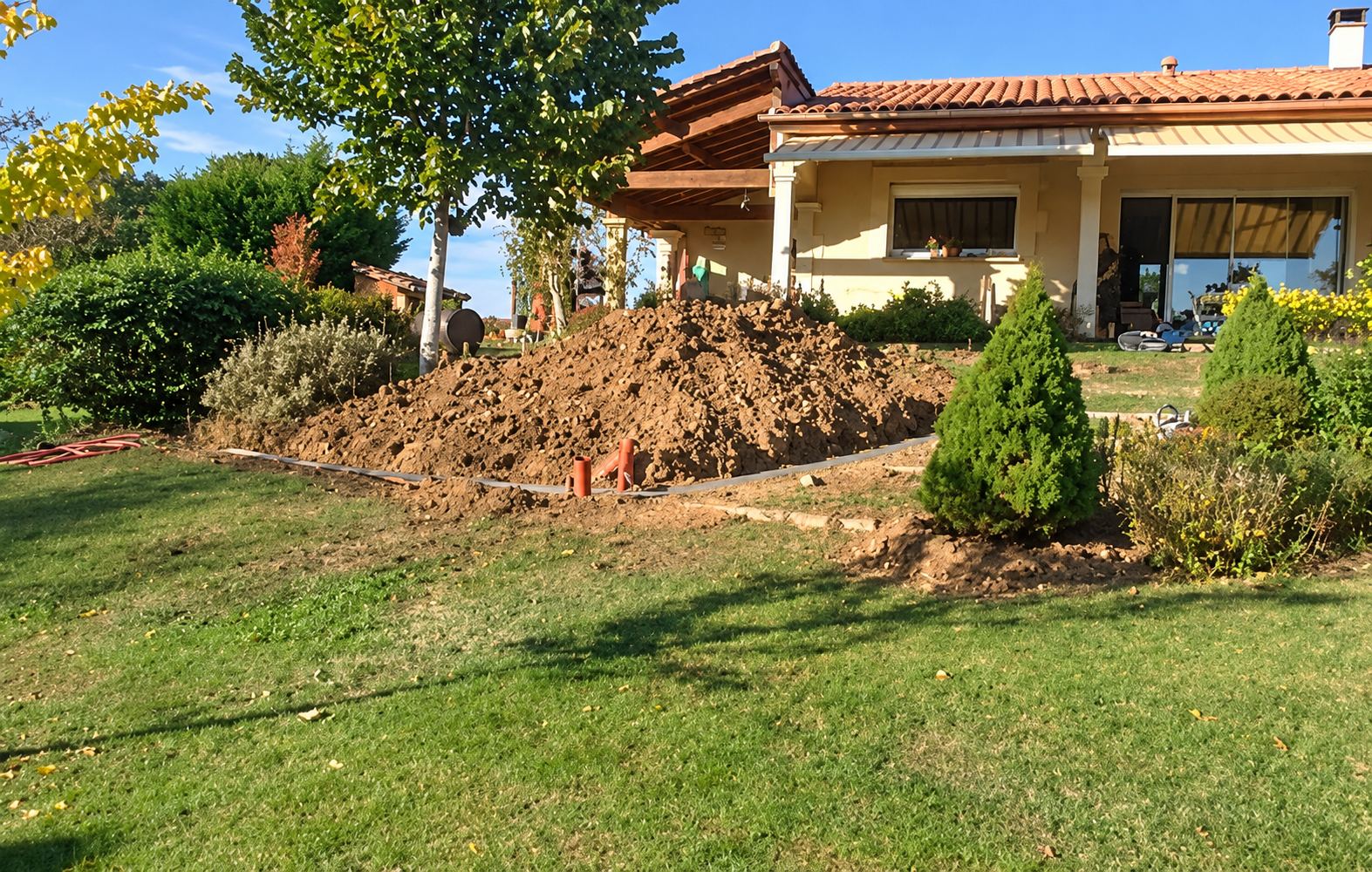 Un grand tas de terre fraîchement creusée repose sur une pelouse devant une maison à toit de tuiles, avec des tuyaux orange qui dépassent du sol.